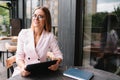 Portrait of young attractive businesswoman examining paperwork in bight light office interior sitting next to the window