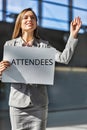 Portrait of young attractive businesswoman standing while holding white board with ATTENDEES signage in arrival area at airport