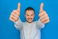 Portrait of young attractive european happy businessman in white shirt with beard showing cool thumbs up two hands on blue