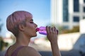 Portrait of young, attractive, gay, heavily makeup man with pink hair and top, drinking water from a bottle on a hot afternoon.