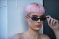 Portrait of young attractive gay man, heavily makeup, pink hair, looking straight ahead over sunglasses, leaning on frame next to