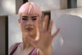 Portrait of young, attractive, gay man, heavily makeup, with pink hair and top, covering the camera as a sign of rejection. LGTBIQ