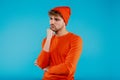 Portrait of young attractive man in orange hat and t-shirt stands in thinkful position on blue background