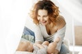 Portrait of young attractive mother laughs and playing with newborn child in comfy light bedroom. Warm mornings with