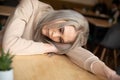 Portrait of young attractive pensive woman with long grey hair with make-up in sweatshirt laying head on hand on table.