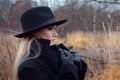 Portrait of young attractive woman in black coat and hat. Lifts collar coat. Autumn landscape, dry grass