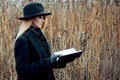 Portrait of young attractive woman in black coat and hat. She's one in a field reading book, autumn landscape, dry grass