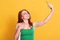 Portrait of young attractive woman making selfie photo with smart phone isolated over yellow background, red haired female in Royalty Free Stock Photo