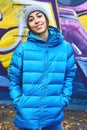 Portrait of young attractive woman wearing in blue down jacket and knitted hat posing against wall painted with graffiti Royalty Free Stock Photo