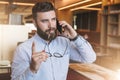 Portrait of young bearded businessman in shirt,holding glasses in hand,lifting his index finger up, and talking on cell Royalty Free Stock Photo