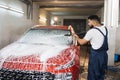 Portrait of young bearded man in working clothes, car wash employee, washing the car windshield Royalty Free Stock Photo