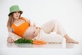 Portrait of young beauteous pregnant woman with long dark hair wearing green bucket hat, lying near bunch of carrots.