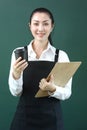 Portrait of Young beautiful Asian female barista wearing apron, holding menu and paper disposable coffee cup on green background, Royalty Free Stock Photo