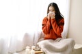Portrait of young beautiful Asian woman sneezing and blowing nose with a tissue sitting home in the chair by the window. Young Royalty Free Stock Photo