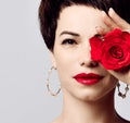 Portrait of young beautiful brunette woman with short hair in round earrings with red lips holding red rose in hand over one eye
