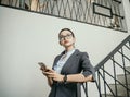 Portrait of young beautiful businesswoman standing in stairs office while holding smartphone and think to get idea