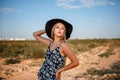 Portrait of a young beautiful caucasian blonde girl in a blue dress with a floral print and a black hat standing on a sandy Royalty Free Stock Photo