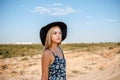 Portrait of a young beautiful caucasian blonde girl in a blue dress with a floral print and a black hat standing on a sandy Royalty Free Stock Photo