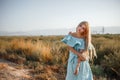 Portrait of a young beautiful caucasian blonde girl in a light blue dress standing on a field with sun-dried grass next to a small Royalty Free Stock Photo