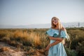 Portrait of a young beautiful caucasian blonde girl in a light blue dress standing on a field with sun-dried grass next to a small Royalty Free Stock Photo