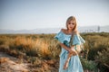 Portrait of a young beautiful caucasian blonde girl in a light blue dress standing on a field with sun-dried grass next to a small Royalty Free Stock Photo