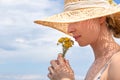 Portrait of young beautiful cheerful woman wearing straw sun hat, smelling small bouquet of yellow wild florets, against