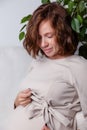 Portrait of young beautiful curly pregnant woman in beige nude dress holding hands on her tummy on background of white