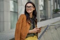 Portrait of a young beautiful and fashionable woman wearing eyeglasses holding her smartphone, looking aside and smiling Royalty Free Stock Photo
