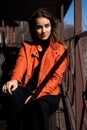 Portrait of young beautiful girl in shadow wearing a orange jacket. Woman sits on the terrace and looking into the Royalty Free Stock Photo