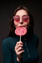 Portrait of a young beautiful stylish girl wearing red drop shaped sunglasses holding a red lollipop and biting it in a Royalty Free Stock Photo