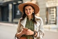 Portrait of a young beautiful stylish woman wearing autumn coat and hat holding her smartphone, looking at camera and Royalty Free Stock Photo
