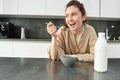 Portrait of young beautiful woman in bathrobe, eating cereals for breakfast, leans on kitchen worktop, looking at her Royalty Free Stock Photo