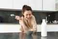 Portrait of young beautiful woman in bathrobe, eating cereals for breakfast, leans on kitchen worktop, looking at her Royalty Free Stock Photo