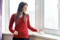 Portrait of young beautiful woman in red sweater drinking coffee. Female standing near window in skyscraper in morning on cloudy Royalty Free Stock Photo