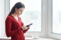 Portrait of young beautiful woman in red sweater reading text on smartphone and drinking coffee. Woman standing near window in Royalty Free Stock Photo