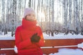 Young beautiful woman in red warm jacket sits on bench in pictorial park with birches and warms hands in winter frosty sunny day Royalty Free Stock Photo