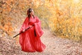 Portrait of young and beautiful woman wearing red chinese warrior costume with black sword, she post using sword among green trees Royalty Free Stock Photo