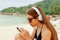 Portrait of a young beauty teenager girl sitting by white sand beach listening to music using headphones and smartphone against on Royalty Free Stock Photo