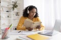 Portrait of young black lady in wireless headphones sitting at desk, using laptop and reading book at home Royalty Free Stock Photo