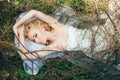 Portrait of young blond bride with veil that lies on earth under the branches and hands up