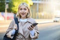 Portrait of young blond woman with backpack, holding mobile phone, standing on street on sunny day, looking surprised Royalty Free Stock Photo