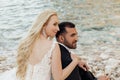 Portrait of young blonde bride in white dress and brunet groom in suit embracing and sitting on sea coast in Italy back Royalty Free Stock Photo