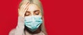 Portrait of young blonde girl having headache, holding hand on head, wearing medical face mask against coronavirus on red. Royalty Free Stock Photo