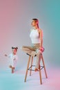 Portrait of a young blonde in a light one sitting on a stool in the studio. A little girl is running around. Mixed blue and pink