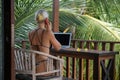 Portrait of young blonde woman freelancer working with laptop on balcony of tropical bungalow with palm trees view Royalty Free Stock Photo