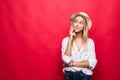 Portrait of young blonde woman wearing in straw hat and touching chin with brooding or dreaming view, isolated over red background Royalty Free Stock Photo
