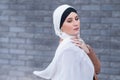 Portrait of a young blue-eyed woman in a hijab against a gray brick wall. A Muslim woman looks at the camera turning