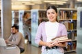 Smiling young woman with stack of books standing in library Royalty Free Stock Photo