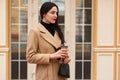 Portrait of young brunette drinking coffee, waiting boyfriend in city. Wearing elegant coat with small black handbag, posing Royalty Free Stock Photo