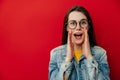 Portrait of young brunette woman screams with excitement, keeps hands near mouth, wears denim jacket and eyeglasses Royalty Free Stock Photo